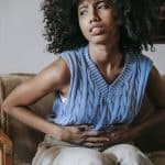 Young African American female sitting on couch at home while having acute pain in stomach